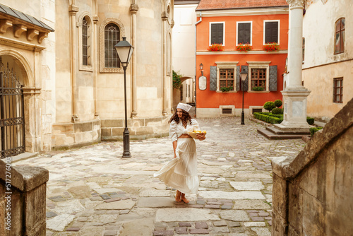 Happy smiling curly woman wearing trendy summer linen shirt, bandana, maxi skirt, holding bowl with lemons, walking in street of European city. Full-length outdoor portrait. Copy, empty space for text