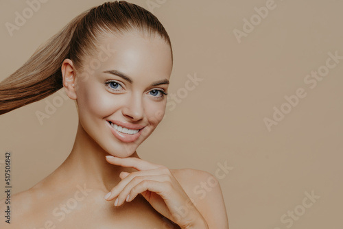 Tranquil undressed woman with healthy perfect skin, dark hair combed in pony tail, enjoys facial treatments, has well cared complexion, stands against beige studio wall, empty space. Skin care concept