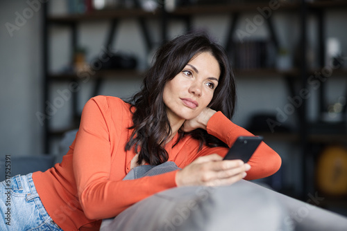 Attractive brunette woman sitting on sofa at home and thinking while text messaging on cell phone. Beautiful female contemplating over sms on mobile phone