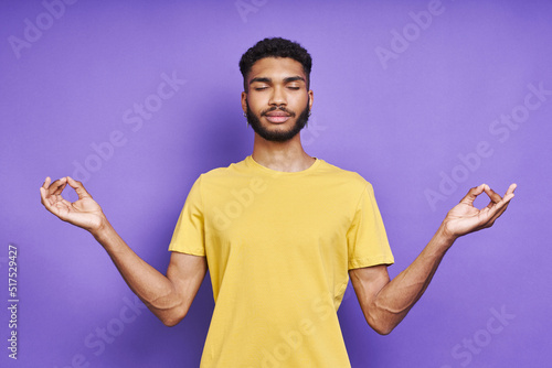 Young African man meditating with his eyes closed while standing against purple background