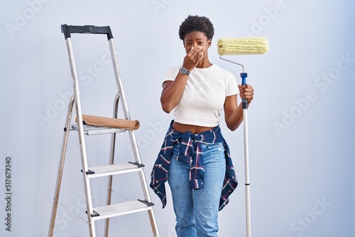 African american woman holding roller painter smelling something stinky and disgusting, intolerable smell, holding breath with fingers on nose. bad smell