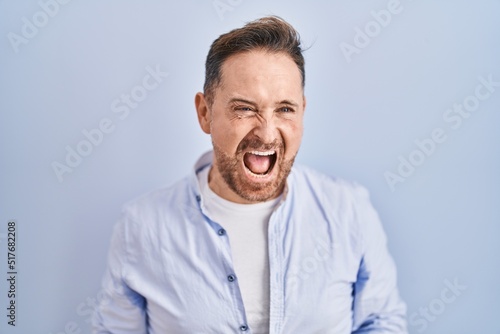 Middle age caucasian man standing over blue background angry and mad screaming frustrated and furious, shouting with anger. rage and aggressive concept.