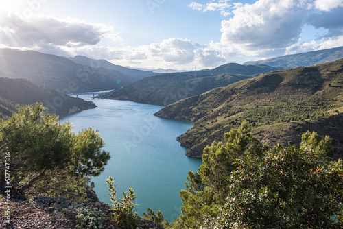 Breath-taking view from the top on green lake surrounded by picturesque mountain alley with trees and bushes in sunny summer day with blue sky and white clouds in background in Spain Andalusia