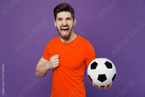 Young expressive excited happy fun fan man he wears orange t-shirt cheer up support football sport team hold in hand soccer ball watch tv live stream scream shout isolated on plain purple background.