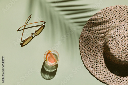 Summer flat lay with straw hat, sunglasses and glass of water with grapefruit slices on green background with palm leaf shadow, sun and sunlight. Vacation, holiday, summer creative minimal concept.