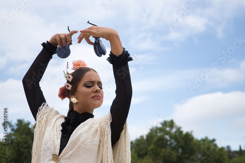 Portrait of young teenage girl in black dance dress, white shawl and pink carnations in her hair, dancing flamenco with castanets in her hands. Concept of flamenco, dance, art, typical Spanish dance.