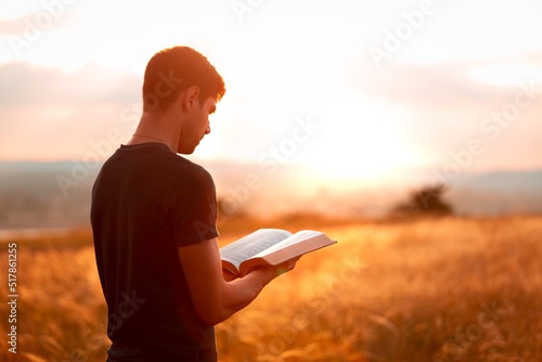 Human praying on the holy bible in a field during beautiful sunset.