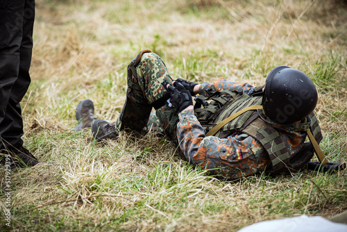a military soldier in a bulletproof vest uniform and a helmet helmet lies on the grass on the grass and puts a jgut turnstile on his leg. a soldier puts a tourniquet on his wounded leg