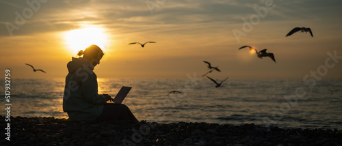 Caucasian woman typing on a laptop on the seashore at sunset. Freelance work. Wide screen. 