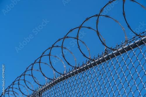 Fence with razor wire with blue sky in the background is shown. Razor wire is made up of high tensile core wire and a punched steel tape with sharp barbs at close intervals uniformly. 
