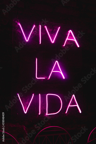 Sign in Spanish with neon light with the text Viva la Vida. Neon lights sign on a black background. bright letters. A pink neon sign lit up a room.