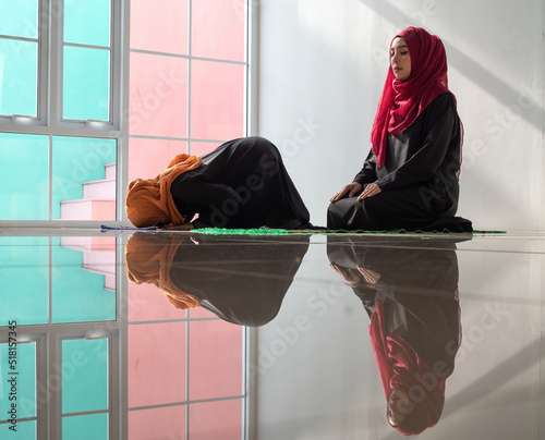 Two Muslim women, one making prostration praying in Sujud pose the other one meditating pose. The shiny floor reflected the reflection of two Muslim lady praying.