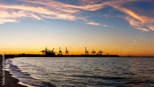 Sunset over harbor in Gdansk with commercial dock and cranes. View from Stogi beach in Baltic sea. Silhouette of industrial port