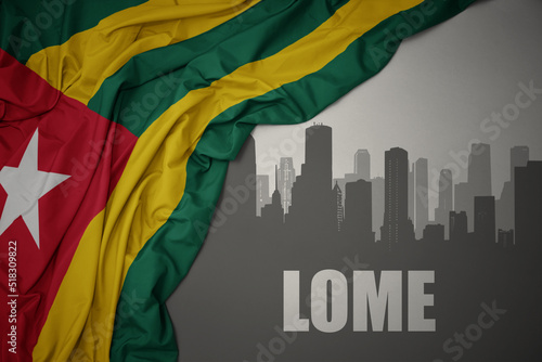 abstract silhouette of the city with text lome near waving colorful national flag of togo on a gray background.