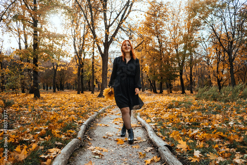 Outdoor autumn portrait of happy smiling plus size red hair woman in dress walking in fall park. Candid portrait of happy curvy woman