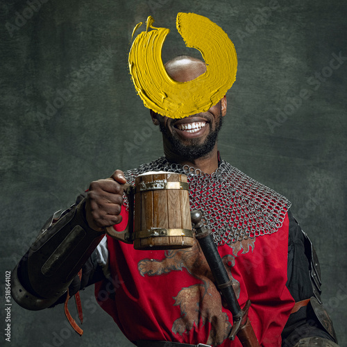 Artwork. Young man as medieval warrior, knight isolated on dark vintage background. Random stroke of yellow paint on his face. Art, creativity, eras comparison concept.