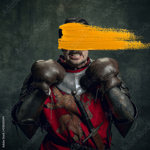 Artwork. Young man as medieval warrior, knight isolated on dark vintage background. Random stroke of yellow paint on his face. Art, creativity, eras comparison concept.