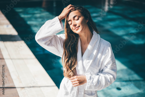 Young woman in white bathrobe walking near swimming pool on background.