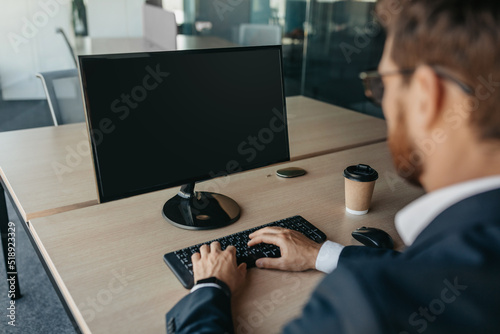 Unrecognizable businessman using computer with empty screen at office, sitting at workplace, offering space for mockup