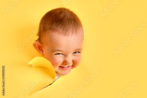 The cunning face of a child looks out of a hole in the studio yellow background. An insidious baby boy peeks through a torn paper background, copy space. Kid age one year