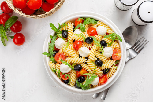 Italian cold pasta salad. Pasta fredda alla caprese. Fusilli, tomato, mozzarella, olive, arugula. Directly above on a white table surface.