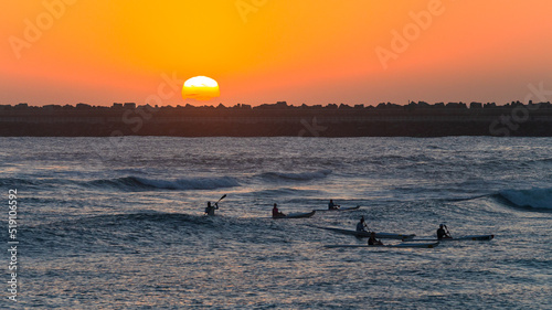 Athlete Paddlers Silhouetted Paddling Surf Ski Crafts Riding Ocean Waves at Sunrise with Harbor Pier and Sky contrasts for a scenic morning landscape.