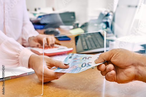 Hand of female social worker taking or giving euro money from client at office workplace with plexiglass separator. Counting grant subsidy rates. Government aid during financial crisis.