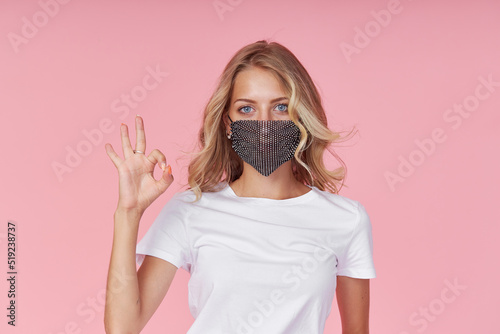A young attractive woman in a white casual t-shirt wears a stylish fashionable protective face mask. Protection against flu and cold diseases during a pandemic. Pink isolated background