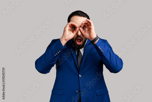 Portrait of astonished surprised bearded man standing raised arms and looking with binoculars gesture, wearing official style suit. Indoor studio shot isolated on gray background.