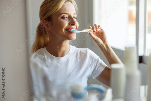 Close up of cheerful young blonde female brushing teeth with toothbrush at mirror. 