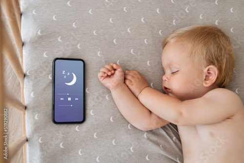 Upper view of baby napping in his bed next to phone playing lullaby in app for baby sleep with child tracker function and parents remote control settings. Maternity and technology. Healthy sleep