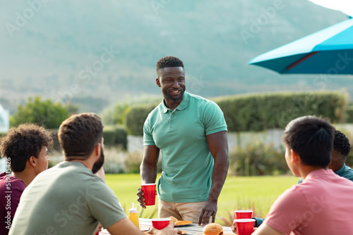 Happy multiracial young man standing and talking with friends while enjoying drinks and food in yard
