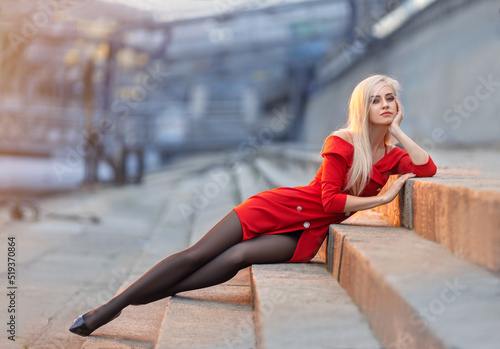Beautiful girl in red dress with perfect legs in pantyhose and shoes with high heels sitting on the street.