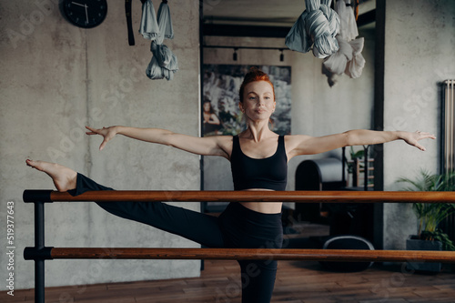 Young attractive woman with perfect body shape stretching on ballet barre during workout in fitness studio