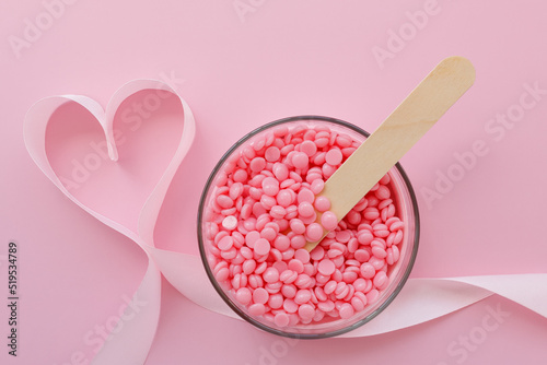 Beautiful granules of pink wax for depilation in a glass bowl, a wooden spatula and a ribbon in the shape of a heart on a pink background. I love waxing. Epilation, depilation, unwanted hair removal.