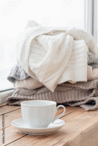 A white cup stands on the windowsill, a pile of warm clothes in the background