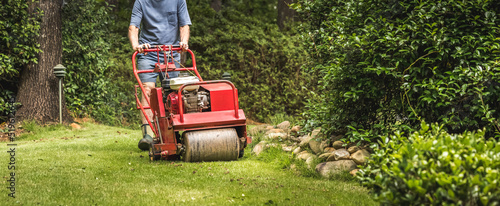 Man using gas powered aerating machine to aerate residential grass yard. Groundskeeper using lawn aeration equipment for turf maintenance.