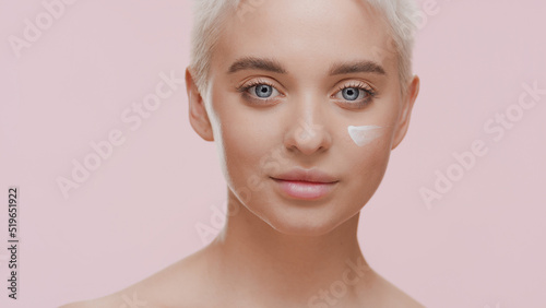 Beautiful young slim light-haired Caucasian woman with white cream on her cheek smiles for the camera looking at it on pale pink background | Face care cream commercial
