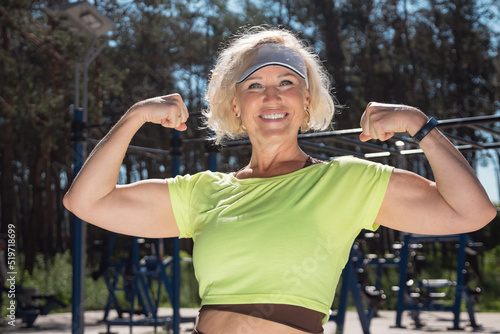 Mature woman shows powerful arm biceps against green park. Middle-aged blonde lady wearing white cap sun visor enjoys healthy life on sports ground