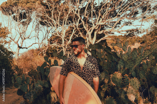Surfer z deską surfingową, idący wybrzeżem na naturalnym tle, drzewa i kaktusy.