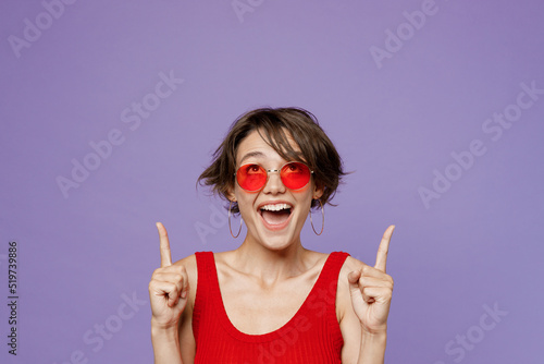 Young shocked amazed happy woman 20s she wear red tank shirt eyeglasses pointing index finger overhead indicate on workspace area copy space mock up isolated on plain purple backround studio portrait