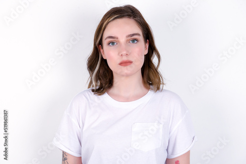 Joyful young caucasian woman wearing white T-shirt over white background looking to the camera, thinking about something. Both arms down, neutral facial expression.