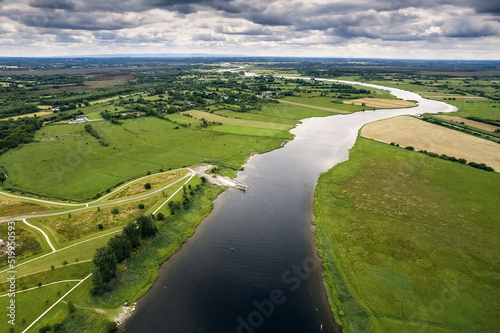 River Shannon flows between green grass fields and agriculture land. Irish landscape. Low cloudy sky. Aerial view. Athlone, Ireland.