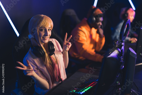 Teenage gamer girl wearing cute ponytails and headset sitting in gaming chair by professional computer smiling looking into camera showing peace signs. High quality photo