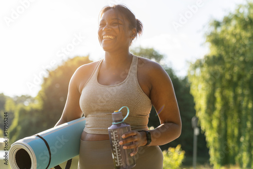 Young African American woman walking with exercise mat and bottle of water through the park in a summer day