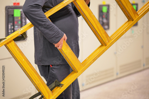 Electrician worker with a yellow wooden staircase. Electrician holds a yellow wooden ladder. Electrician bears a wooden step-ladder in a room on electrical cabinets background