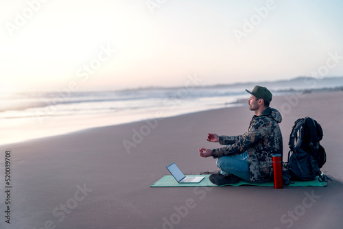 mature digital nomad sitting on the beach meditating with his laptop on the shore of the beach