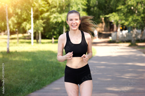 happy fit fitness athletic girl runner running, jogging, young beautiful woman is smiling training outdoors at summer sunny day in the morning in black top and shorts looking at camera
