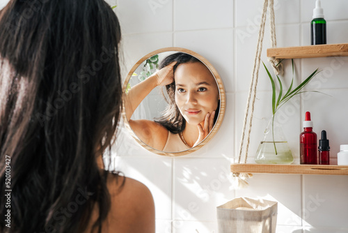 Beautiful hispanic woman looking in the mirror in white eco friendly bathroom. Wooden shleves and reusable cosmetics bottles. Wellnes concept
