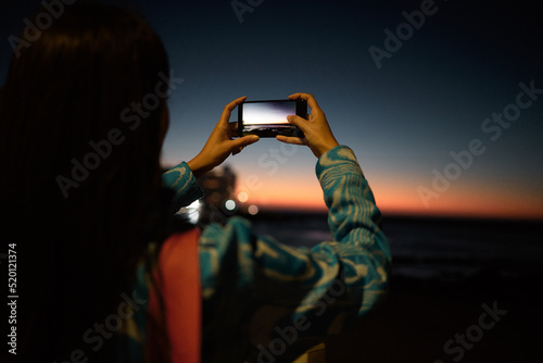Trendy tourist taking photo on her phone of the ocean with beautiful sunset on the horizon at night outdoors. Influencer or vlogger taking picture of nature sea view to post or share on social media